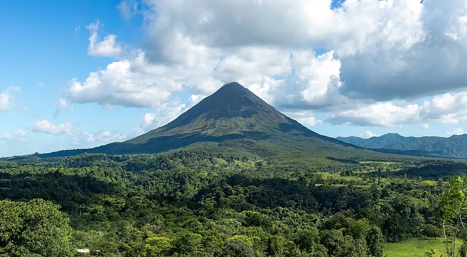 The forested cone of Arenal volcano rising above lowland forest and pasture under scattered cloud.