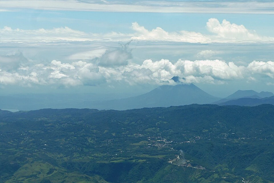 Arenal volcano seen from the air, its cone rising through haze beyond forested ridges, farmland and a small town, with banks of white cloud along the horizon.