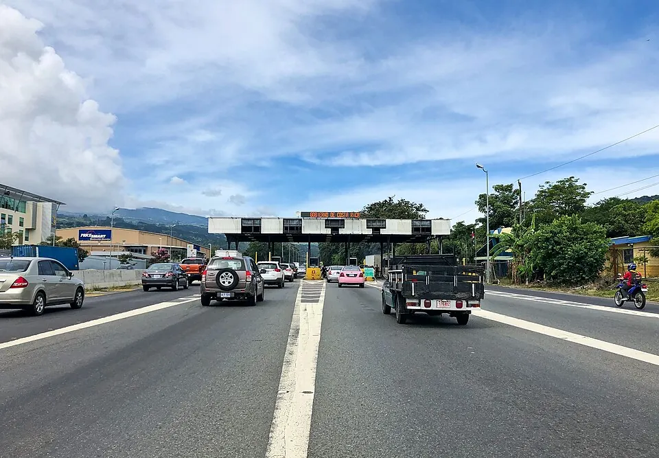 Cars, a flatbed truck and a motorcycle approaching a toll gantry signed Gobierno de Costa Rica on a divided highway.