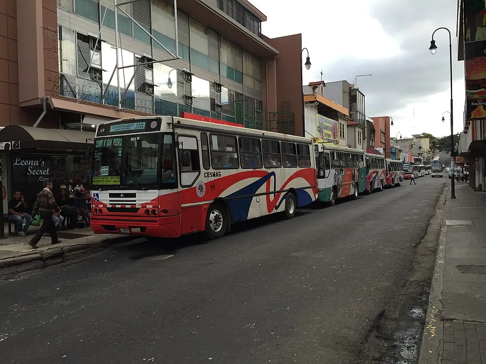 Several public buses queued nose to tail at a kerbside stop on a street in downtown San José, with passengers waiting under a shelter.