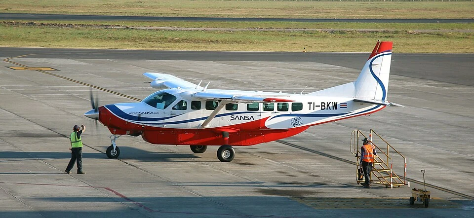 A red and white Sansa Cessna 208B Grand Caravan single-engine turboprop parked on the apron at Liberia airport, with two ground crew beside it.