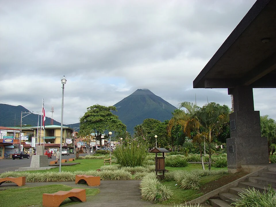 A park in La Fortuna with benches, palms and a Costa Rican flag, shops along the street behind it, and the cone of Arenal volcano rising beyond.