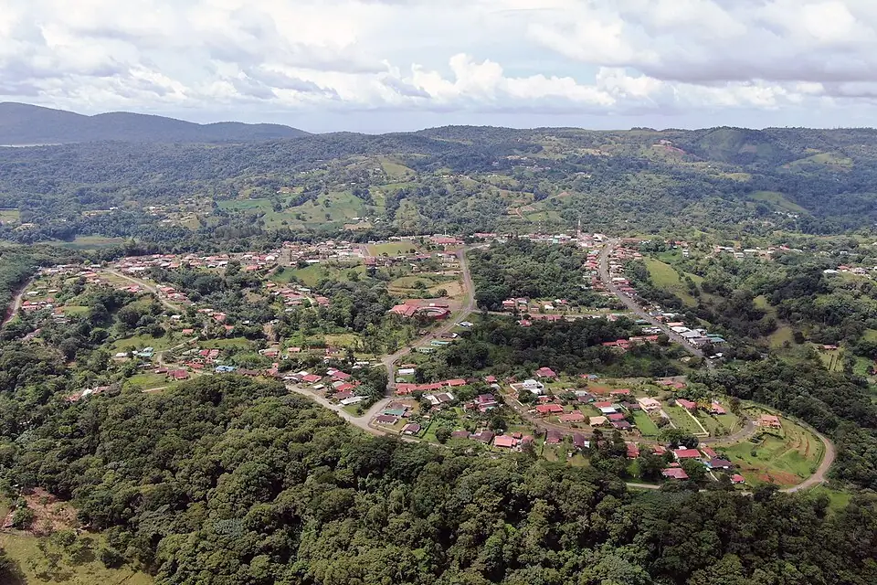 Aerial view of Nuevo Arenal, a small town of red-roofed houses on curving streets among green hills, with Lake Arenal visible in the distance.