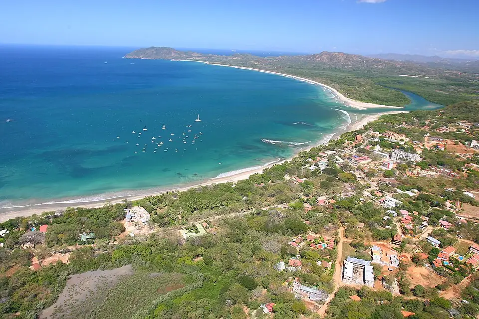 Aerial view of Tamarindo: the town's low buildings behind a long curving beach, sailing boats moored in turquoise water and an estuary running inland.