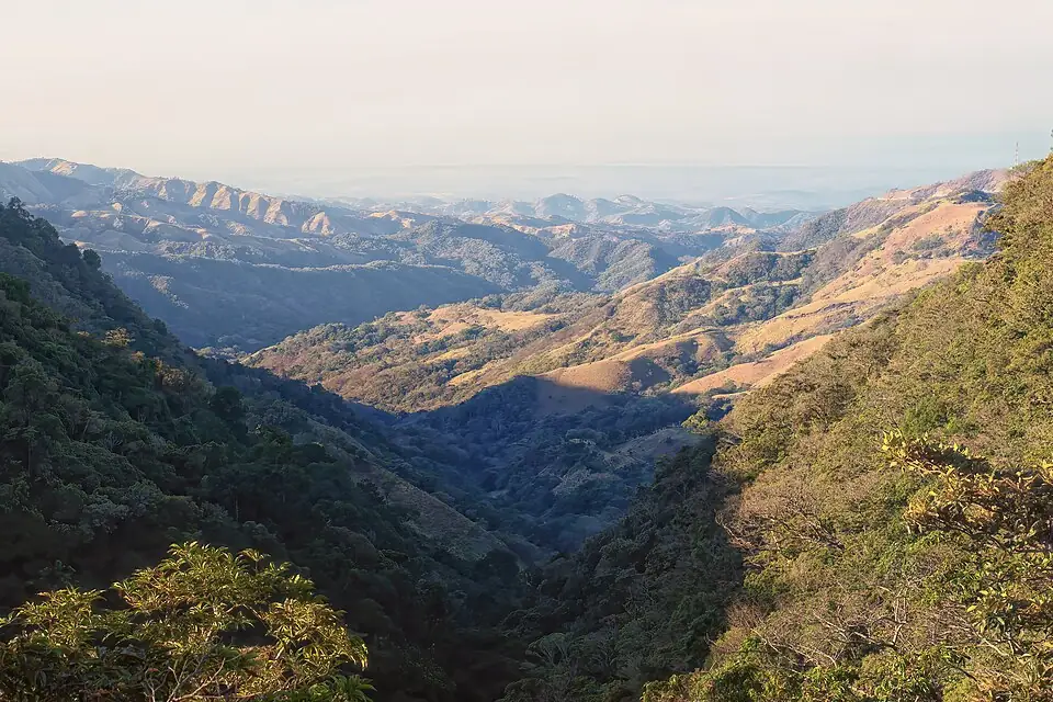 Forested ridges and pasture-covered hills falling away in layers toward a hazy lowland horizon.