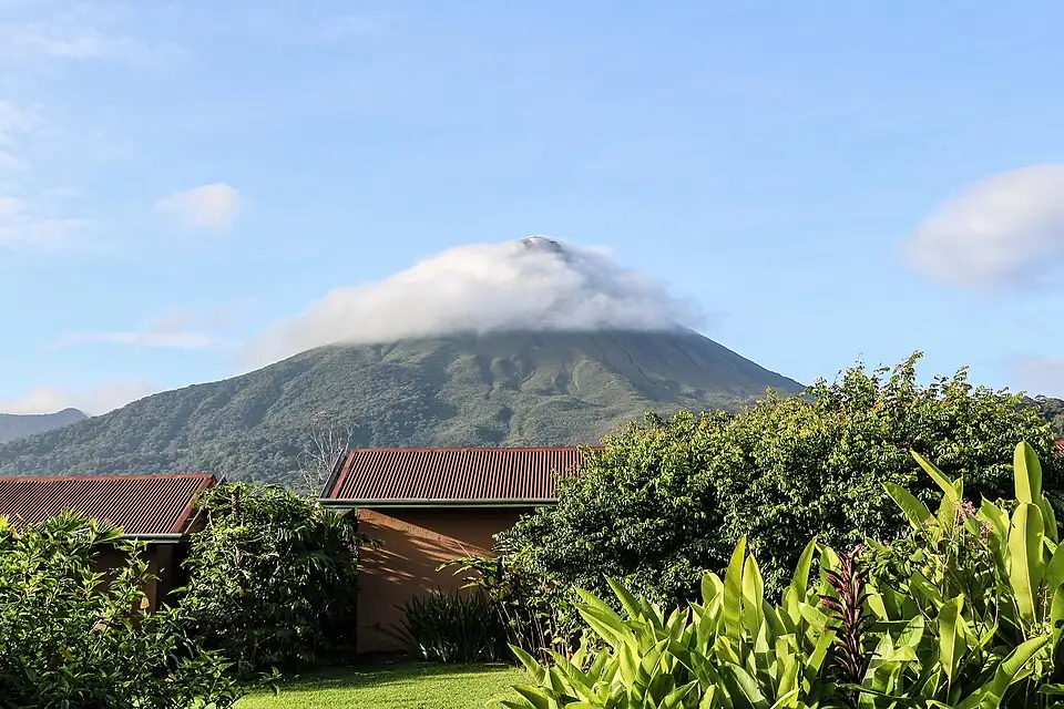 A symmetrical volcanic cone with a band of cloud across its summit, rising behind low red-roofed buildings and garden planting.