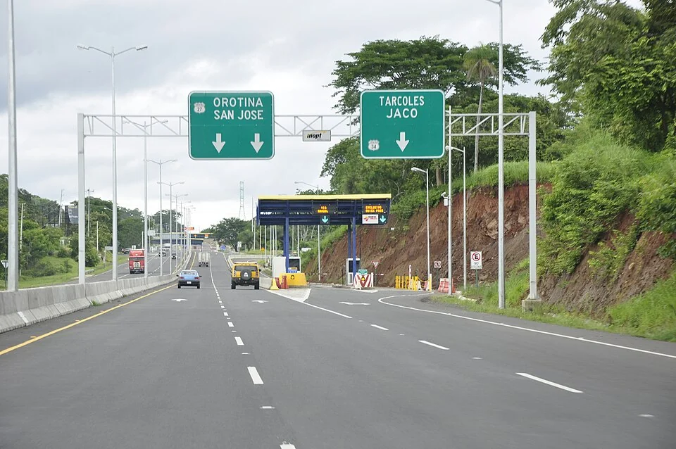 A section of Costa Rica's Ruta 27, with overhead green signs reading Orotina / San José on the left and Tárcoles / Jacó on the right, and a toll plaza ahead on the shoulder.