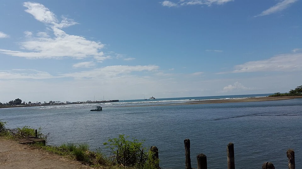 The Quepos waterfront seen across the river mouth, with the town and moored boats on the far shore and the Pacific beyond.