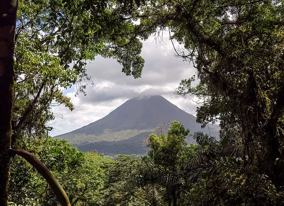 Arenal volcano seen through a gap in dense rainforest canopy, its summit under cloud.