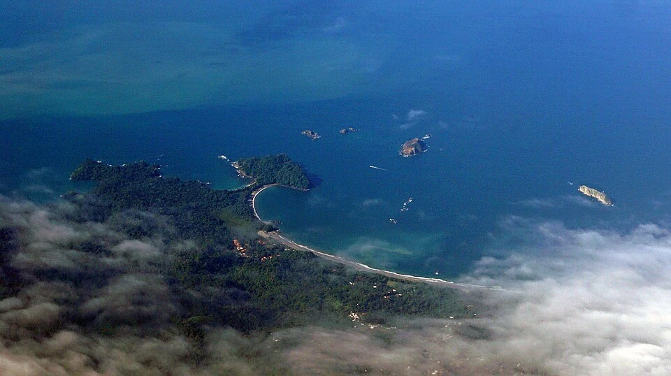 Aerial view of the Manuel Antonio coast: a curving white beach, the forested point beyond it, offshore islets, and buildings scattered across the hillside behind the shore.