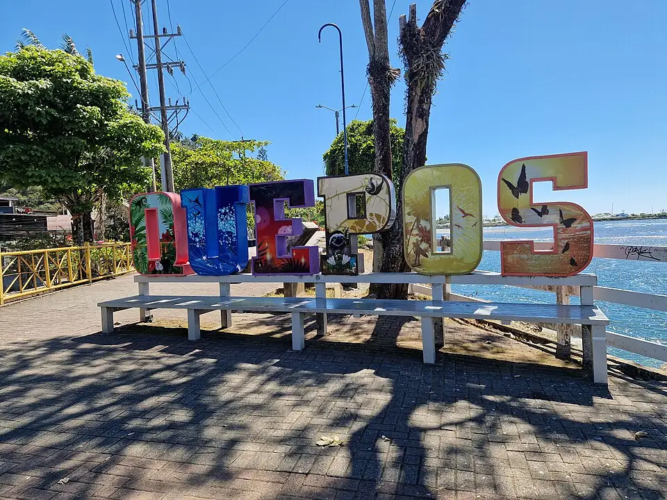 Painted freestanding letters spelling QUEPOS on the seafront walkway in Quepos, Costa Rica, with benches in front and the water behind.