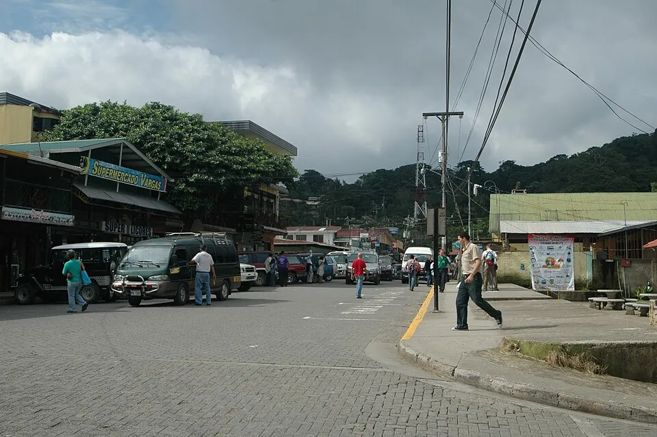 The main shopping street in Monteverde, Costa Rica, with minibuses and pickups parked along it, shopfronts on both sides and forested hills rising behind.