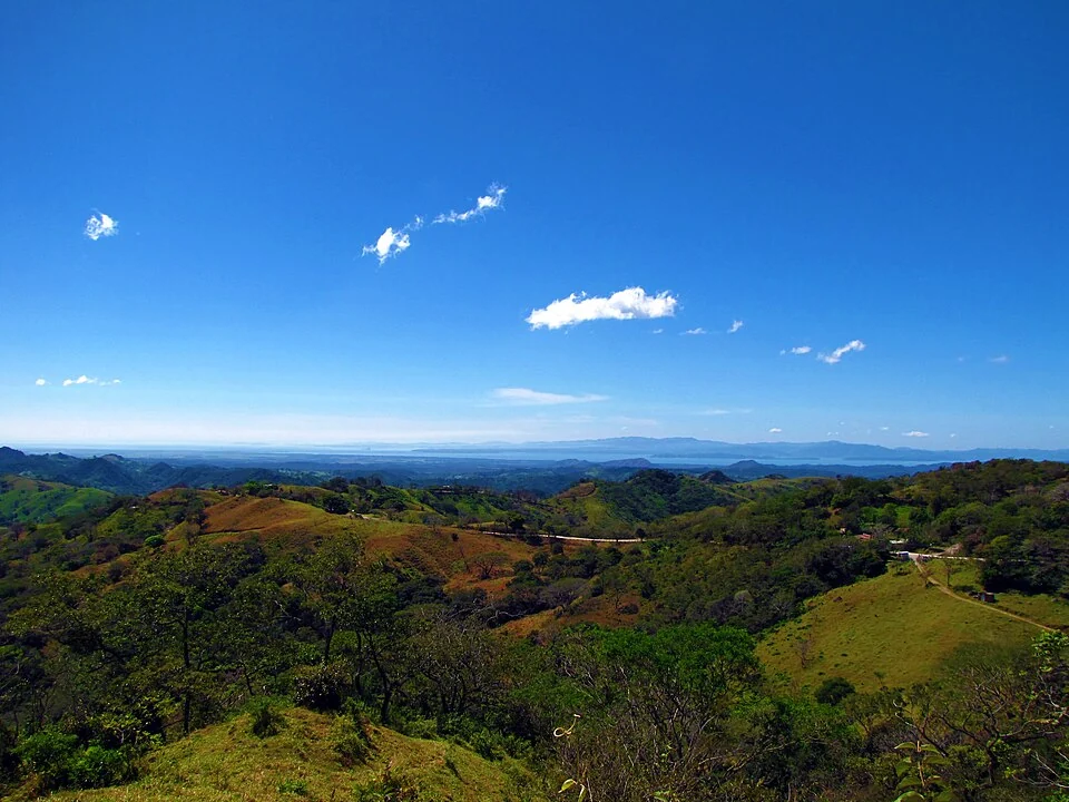 The road to Monteverde crossing green and tan hills, with a few farm buildings along it and the Pacific lowlands and sea in the distance under a blue sky.