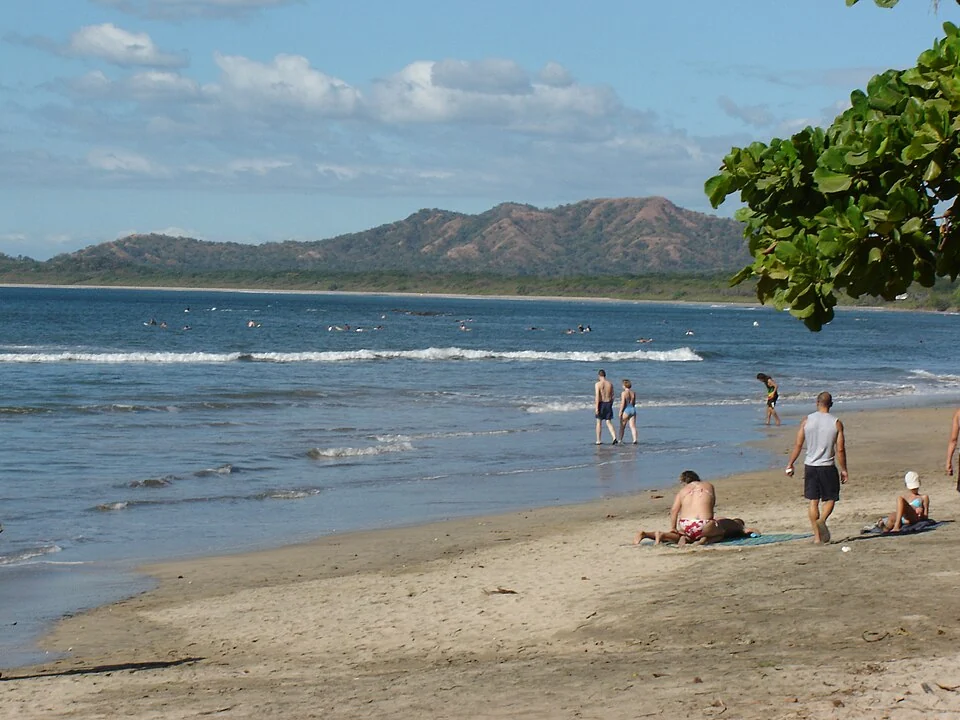 Playa Tamarindo on a clear day, with people on the sand, surfers waiting offshore and dry brown hills across the bay.