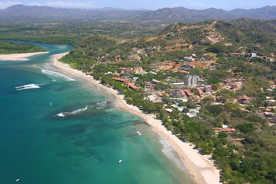 Aerial view of Tamarindo: the beach curving north to the river mouth, the built-up town packed behind it and dry hills inland.